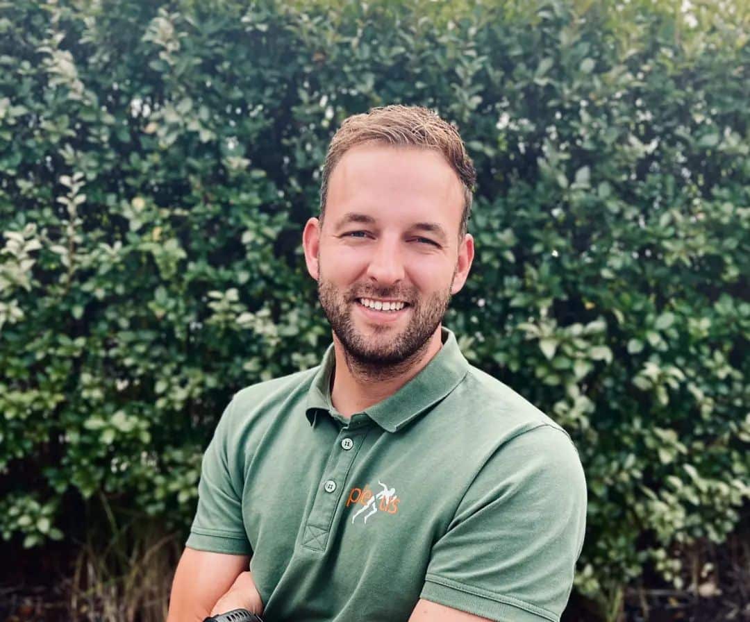 Man in groene polo met logo, lachend buiten met groene achtergrond.